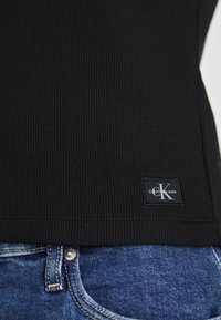 Schwarzes, geripptes Tanktop mit einem eingenähten Calvin Klein Jeans-Label; kombiniert mit blauen Denim-Jeans mit Metallknopf und sichtbaren Nähten.