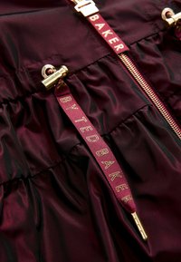 Veste en satin bordeaux avec texture plissée, fermeture éclair dorée, et cordon de serrage avec "TED BAKER" en lettres dorées.