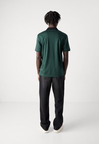 Zelená polo košeľa s krátkym rukávom a čiernym golierom, kombinovaná s čiernymi denimovými džínami. Tkanina pôsobí hladko a má uvoľnený strih.