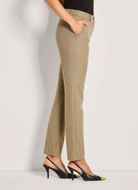 Frau in beigen, gemusterten, geraden Hosen und schwarzen Slingback-Heels mit goldenen spitzen Zehen, Hand in der Tasche, seitlich stehend.