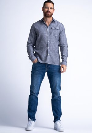 Chemise boutonnière gris clair avec une poche poitrine, associée à un jean bleu et des baskets blanches. La chemise a un tissu texturé et des manches longues.