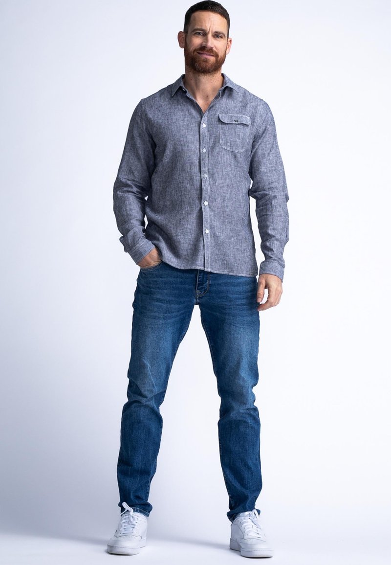 Chemise boutonnière gris clair avec une poche poitrine, associée à un jean bleu et des baskets blanches. La chemise a un tissu texturé et des manches longues.