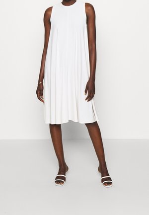 Ärmelloses weißes knielanges A-Linien-Kleid, getragen mit weißen offenen Sandalen mit Absatz an einer stehenden Person.