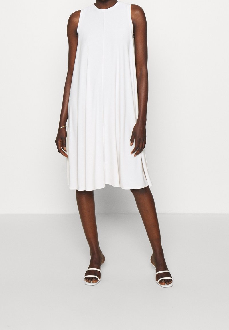 Robe blanche sans manches, longueur genou, coupe trapèze, portée avec des sandales blanches à talons ouvertes, sur une personne debout.