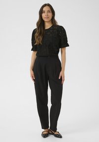 Blouse noire brodée avec des manches bouffantes portée avec un pantalon noir taille haute et des ballerines noires avec des clous décoratifs.