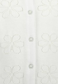 Vit kofta med blommönstrad spets, med glänsande knappar och ribbad halsringning och placket. Mjuk stickad textur.