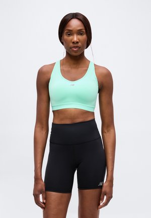Frau steht frontal, trägt einen mintgrünen Sport-BH und hoch taillierte schwarze Sportshorts vor einfachem Hintergrund.