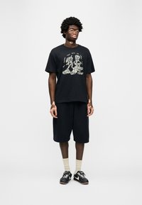 Μαύρο βαμβακερό μπλουζάκι με γραφικό σχέδιο που απεικονίζει περιγραμμένες φιγούρες, συνδυασμένο με μαύρο σορτς και μαύρα αθλητικά παπούτσια με λευκές λεπτομέρειες.