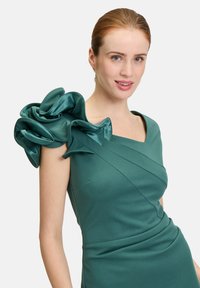 Robe teal avec un corsage ajusté et un accent en satin froncé sur l'épaule gauche. La robe présente une couture diagonale pour ajouter de la texture.