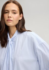 Light blue striped blouse with a tie-neck, featuring vertical stripes, a relaxed fit, and soft fabric texture. Close-up view of the neckline.