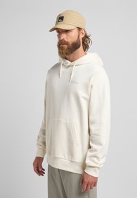 Cremfarbener Kapuzenpullover mit einer Fronttasche und einem kleinen Logo auf der Brust. Weicher Stoff mit gerippten Bündchen und Saum.