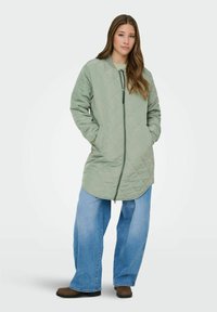 Rūtiņots garš mētelis blāvā zaļā neilona krāsā ar italiņu, sānu kabatām un nedaudz izliekta apakšmalu, kombinēts ar platām zilām džinsu biksēm.