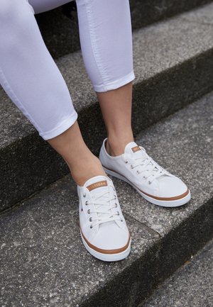 Personne portant des baskets blanches avec des bordures marron et un pantalon blanc assise sur des marches en béton à l'extérieur.
