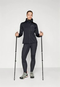 Mladá žena v černém sportovním oblečení držící turistické hole, obutá v šedých a růžových trekových botách, stojící na bílém pozadí.