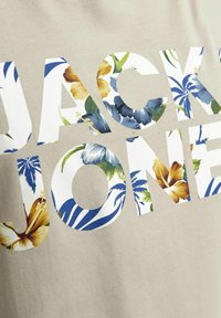 Beige bomull T-skjorte med store blomsterdekorert bokstaver som staver "JACK & JONES" i blå, gul og grønn, med en glatt tekstur.