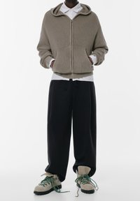 Sweat à capuche zippé vert olive texturé avec poches avant, superposé sur une chemise blanche à col. Pantalon ample noir et chaussures beiges à lacets.