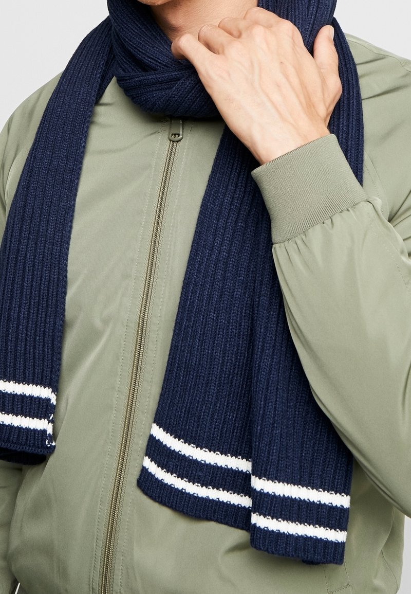 Homme portant une veste zippée vert olive et une écharpe côtelée bleu marine à rayures blanches, tenant l'écharpe près du cou.