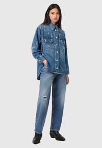 Naine seisab käed taskutes, kandes kaunistatud suure suurusega teksasärki, pleekinud rebenenud teksapükse ja musti pahkluu-saapaid heledal taustal.