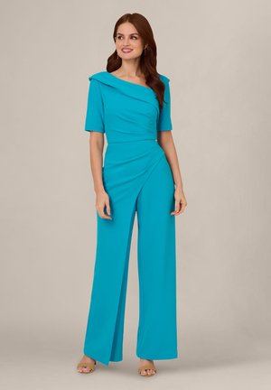 Kobieta z długimi brązowymi włosami ubrana w turkusowy kombinezon o szerokich nogawkach, z asymetrycznym dekoltem i krótkimi rękawami, stojąca na neutralnym tle.