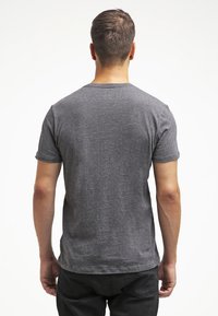 Uomo rivolto verso l'uscita, che indossa una semplice t-shirt grigia a maniche corte e pantaloni neri, su uno sfondo bianco.