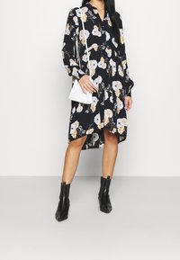 Květinové černé šaty s dlouhými rukávy a límcem; bílá crossbody taška; černé kotníkové boty; přiléhavý korzet, volná sukně.