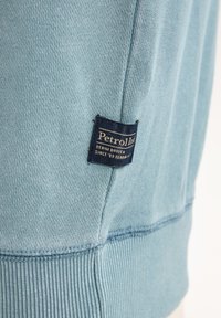 Felpa blu chiaro realizzata in tessuto lavorato a maglia con texture. Presenta un'etichetta blu scuro con testo bianco sul lato. Bordo e orlo a costine.