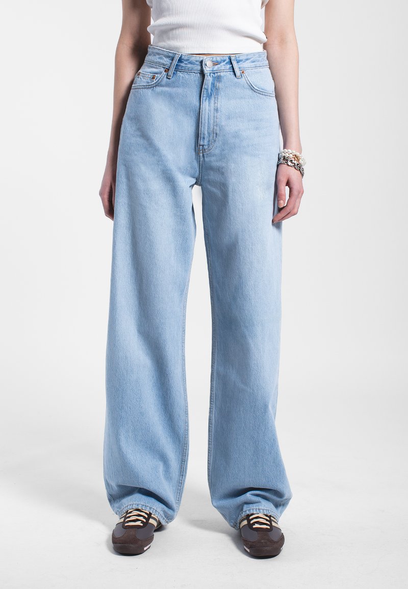 Lichtblauwe broek met ruime pijpen, gemaakt van denim, met een hoge taille, zichtbare stiksels en een relaxte fit, gecombineerd met donkere sneakers.