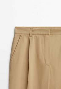 Pantalon beige avec passants pour ceinture, poches latérales et fermeture avant dissimulée, présenté sur fond blanc.