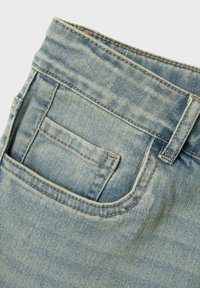 Tela de mezclilla de un azul claro con un bolsillo trasero decorativo, que presenta detalles de costura y una textura suave, parcialmente doblada en el borde superior.