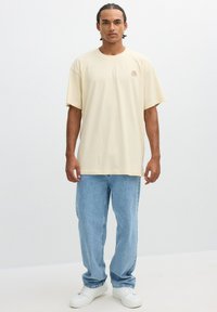 Camiseta de algodón color beige con un corte relajado, mangas cortas y un pequeño diseño de rosa bordado en el lado izquierdo del pecho. Combinada con unos jeans de mezclilla azul claro.