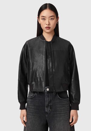 Jeune femme aux cheveux lisses et foncés portant une veste en cuir noire courte sur un haut noir et un jean taille haute foncé.