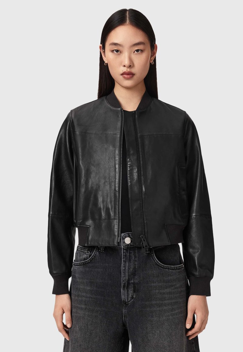 Junge Frau mit glatten dunklen Haaren trägt eine schwarze, kurze Lederjacke über einem schwarzen Oberteil und hoch taillierte dunkle Jeans.