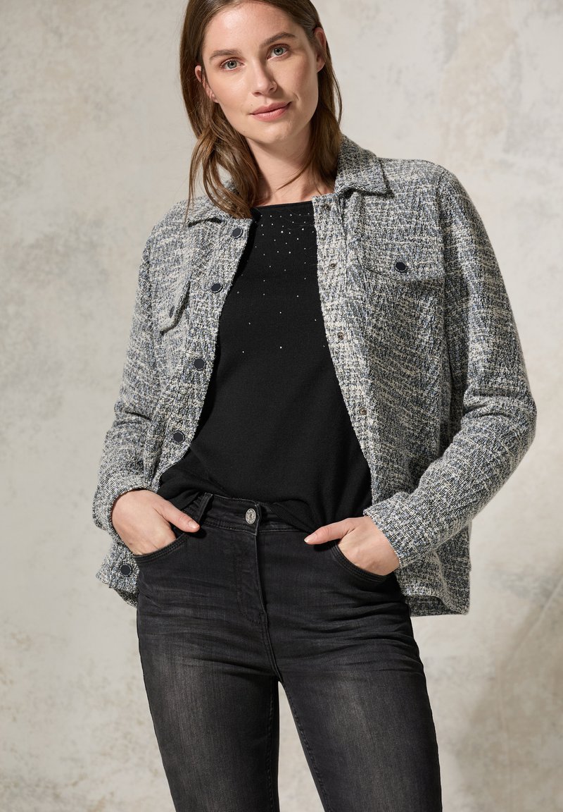 Grau karierter Blazer mit kragenformigem Design, vorderen Taschen und schwarzen Knöpfen, kombiniert mit einem schwarzen Oberteil mit feinen Verzierungen. Schwarze Jeans.