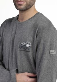 Camisa de manga larga gris hecha de tejido suave, con un diseño impreso de vehículo todoterreno y una etiqueta en la manga izquierda.