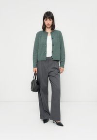 Žena v zeleném button-up svetru, bílém topu, šedých širokých kalhotách, černých špičatých podpatcích, držící černou kabelku, stojící na bílém pozadí.