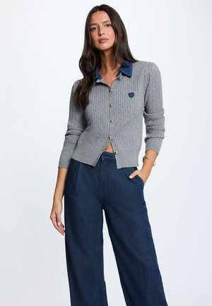Donna con lunghi capelli scuri che indossa un cardigan grigio a coste con colletto blu e pantaloni a gamba larga blu scuro, in piedi con una mano in tasca.