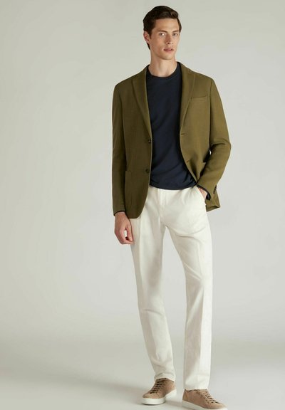 Blazer olive avec un bouton et une poche, t-shirt bleu marine, pantalon blanc et baskets beiges à lacets. Texture lisse dans l'ensemble.