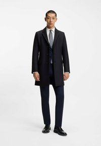 Cappotto in lana navy con vestibilità classica, caratterizzato da un colletto a risvolto, bottoni frontali e tasche laterali, indossato sopra un abito scuro e una cravatta.