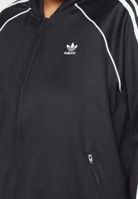 Mustat vetoketjulliset takki, joka on valmistettu sileästä kankaasta. Takissa on valkoiset raidat hartioilla ja pieni Adidas-logo rinnassa.