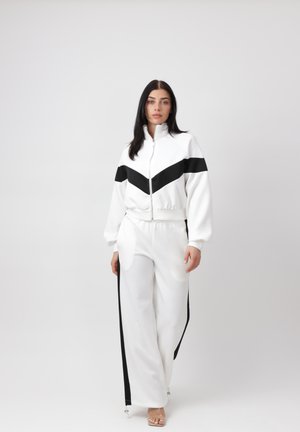 Kvinde stående iført en hvid træningsdragt med sort chevron og sidelister, lynlåset jakke og bukser med vide ben, på en ensartet lys baggrund.