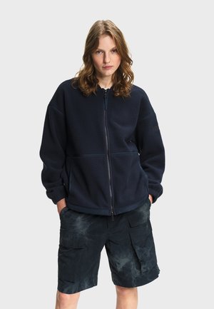 Młoda osoba o falowanych włosach ma na sobie granatową polarową kurtkę zapinaną na suwak i ciemne, wzorzyste szorty typu cargo, stojąc z rękami w kieszeniach.