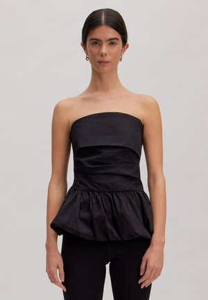 Vrouw met donker haar draagt een strapless zwart topje met een aansluitend lijfje en geplooid peplum, gecombineerd met zwarte broek, tegen een effen achtergrond.