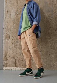 Jeansjacka över en grön t-shirt, tillsammans med beige cargobyxor med fickor och Nike-logga, kompletterat med gröna och svarta sneakers.