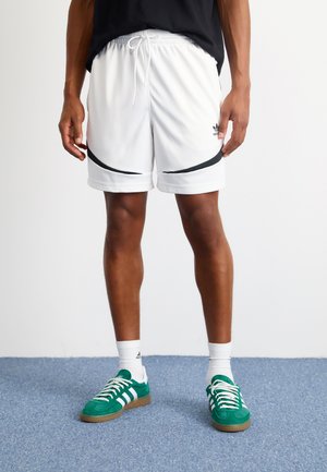 Pantaloncini sportivi bianchi con un accento curvo nero, vita elastica e lacci. Abbinati a sneakers in suede verde e calzini bianchi.