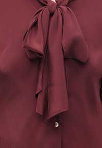 Burgunderbluse aus einem sanften, fließenden Stoff. Sie hat eine große, geknotete Schleife am Ausschnitt und einen einzelnen Knopfverschluss am Kragen.