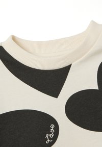 Sweatshirt crème avec de grands motifs de cœurs noirs, col côtelé et un subtil détail textuel "Love" en cursive dans le coin inférieur droit.