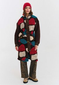 Žena oblečená v farebnom patchwork fleece kabáte, červenej pletenej čiapke, nohaviciach s leopardím vzorom a béžových turistických topánkach, stojí na pozadí so jednoduchým vzorom.