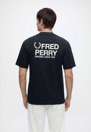 Hombre con camiseta negra de Fred Perry con logo blanco y texto en la espalda, combinada con pantalones blancos, de espaldas sobre un fondo liso.