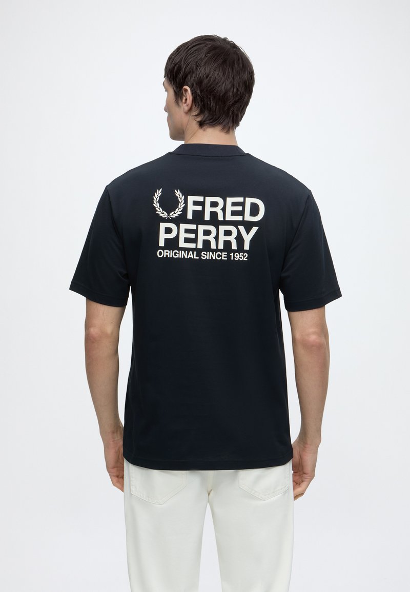 Άνδρας φοράει μαύρο μπλουζάκι Fred Perry με λευκό λογότυπο και κείμενο στην πλάτη, σε συνδυασμό με λευκό παντελόνι, γυρισμένος με την πλάτη σε απλό φόντο.