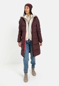 Bordeaux puffer jas met capuchon en witte fleece voering, gelaagd over een lichtbeige top, gecombineerd met blauwe jeans en beige laarzen.